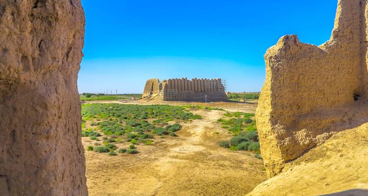 Earthen desert fortifications rise from a scrub landscape framed by fractured walls and vivid blue sky.
