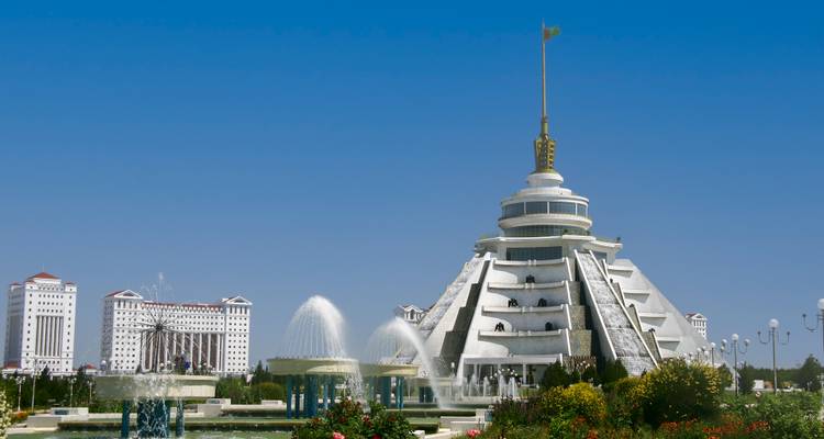 Futuristic white marble building with fountains and gold spire dominates the skyline under a clear blue sky.