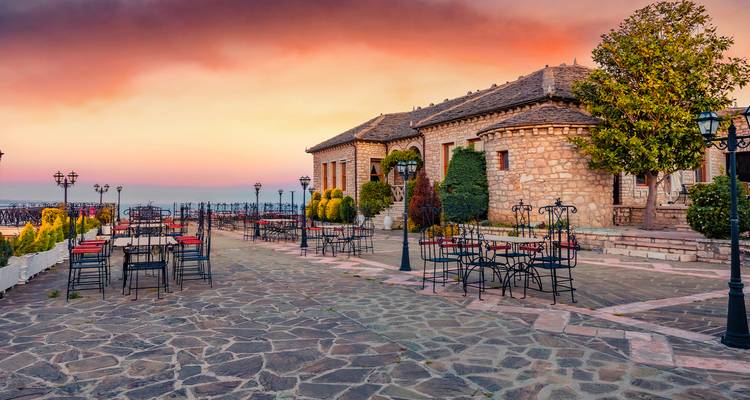 Terrasse pavée de pierres bordée de tables en fer forgé donnant sur un bâtiment historique en pierre au coucher de soleil éclatant.