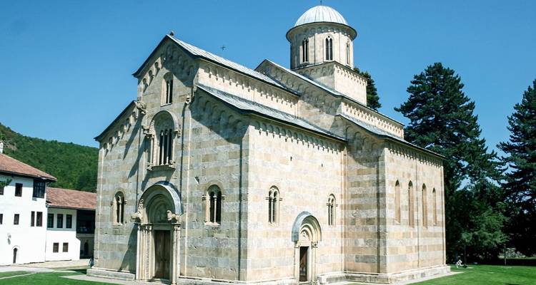 Well-preserved medieval stone monastery with a domed roof set on a green lawn under a bright blue sky.