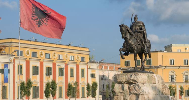 Equestrian statue of a national hero and a large Albanian flag dominating a sunny city square lined by colourful buildings.