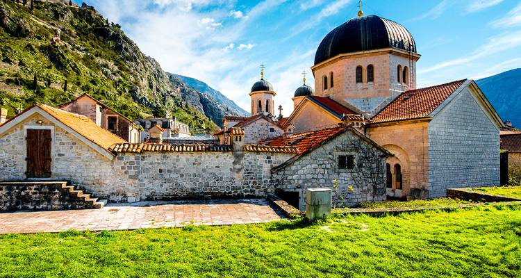 In Kotor gelegene, aus Stein erbaute orthodoxe Kirche mit Kuppeldächern vor grünen Bergen und blauem Himmel.