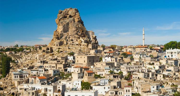 Stenen huizen opgestapeld aan de voet van het imposante Ortahisar rotskasteelonder een kristalheldere blauwe Cappadociëhemel.