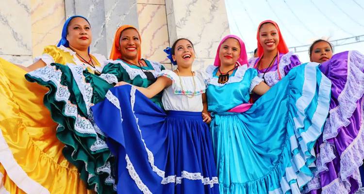 Seis mujeres vistiendo coloridos trajes tradicionales y sonriendo ampliamente en formación de danza folclórica.