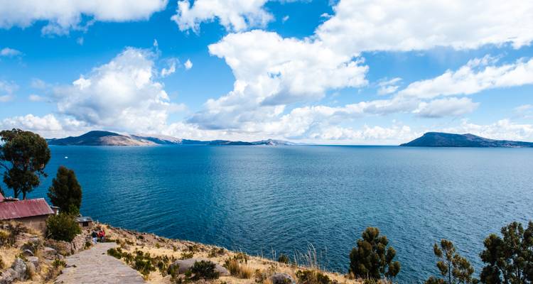 Weite Panoramaaussicht auf einen großen blauen See unter einem Himmel voller heller weißer Wolken und entfernter Hügel.