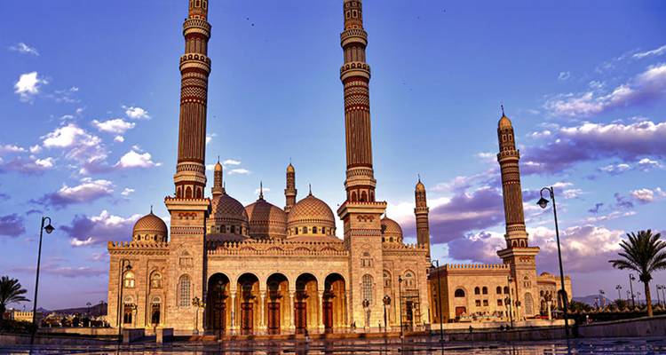 Una gran mezquita con múltiples minaretes y una ornamentada cantería de ricos diseños se baña en la cálida luz del atardecer bajo un cielo púrpura-azul.
