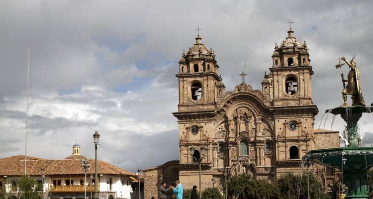 Les tours d'église baroque en pierre historique s'élèvent au-dessus de la Plaza de Cusco avec les habitants et les visiteurs rassemblés en bas sous un ciel nuageux.