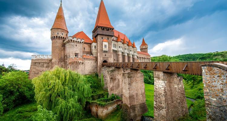 Medieval Corvin Castle with red-tiled towers and a wooden bridge rising above lush green hills under a dramatic cloudy sky.