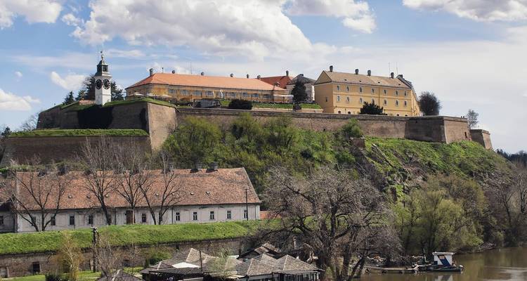 Historic Petrovaradin Fortress with clocktower and ochre buildings perched on a green bluff above a river.