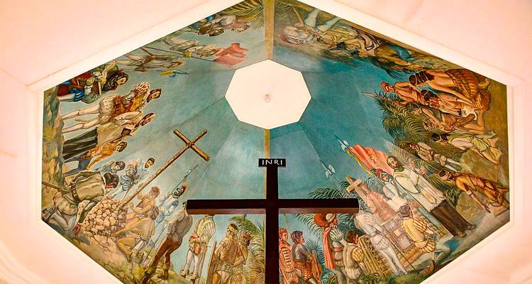 Une fresque murale octogonale dépeignant des scènes religieuses historiques avec une croix en bois au centre.