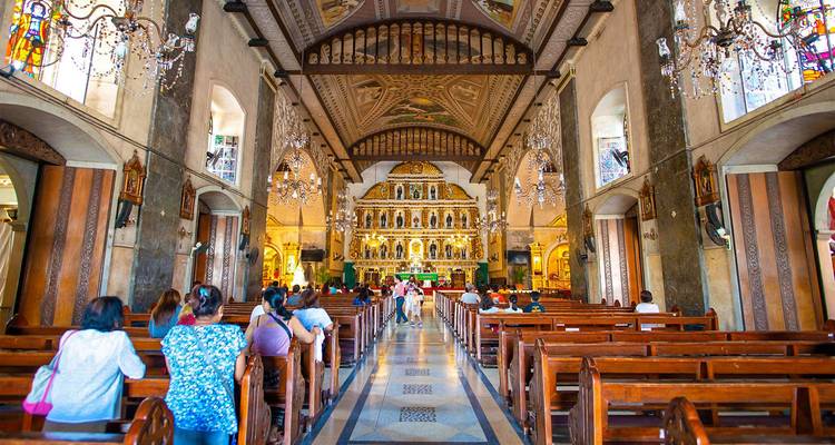 L'intérieur orné d'une église philippine avec des lustres et des fidèles assis le long d'une longue allée centrale.
