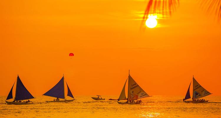 Des voiliers en silhouette glissent sur une eau dorée sous un coucher de soleil orange flamboyant.