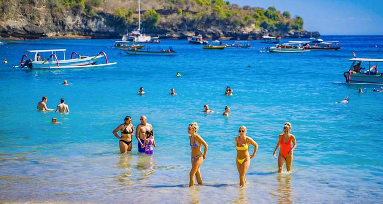 Les baigneurs nagent et pataugent dans une eau turquoise éclatante tandis que les bateaux mouillent dans une baie abritée.