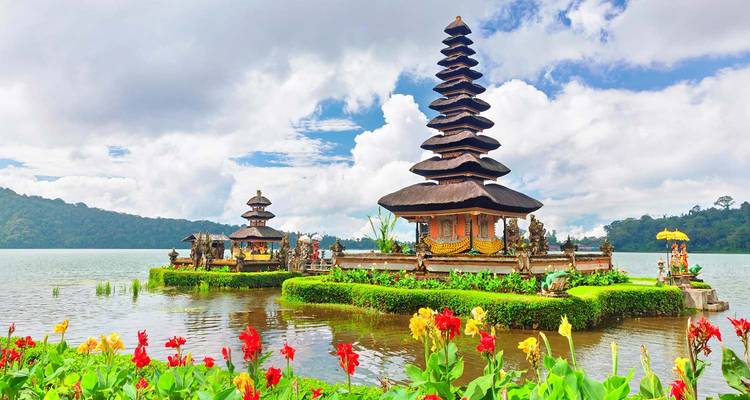 Un temple hindou balinais à étages est situé sur de petites îles entourées d'un lac calme avec des fleurs colorées au premier plan et des collines boisées en arrière-plan.