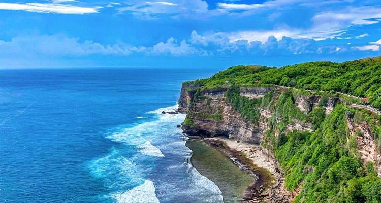 Des falaises spectaculaires recouvertes de végétation verte plongent dans les vagues océaniques d'un bleu turquoise éclatant le long d'une côte accidentée sous un ciel bleu dégagé.