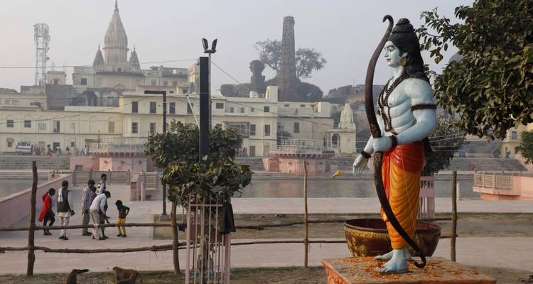 Statue of Lord Rama with a bow and buildings in the background near a river.