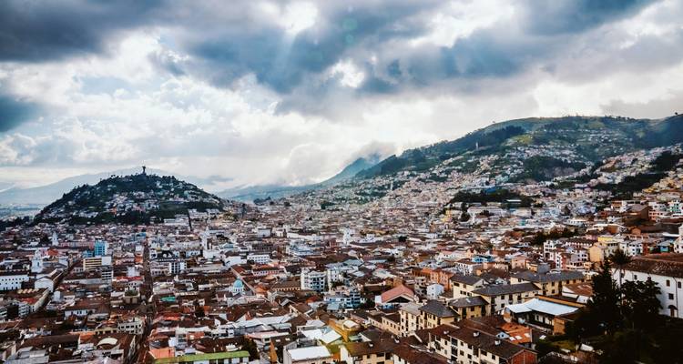 Vista expansiva sobre los techos de Quito bajo nubes dramáticas con la luz del sol filtrándose en la ciudad.