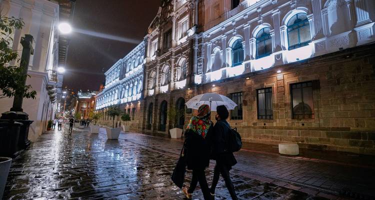 Escena callejera nocturna en el centro histórico de Quito con dos personas compartiendo un paraguas sobre adoquines mojados.