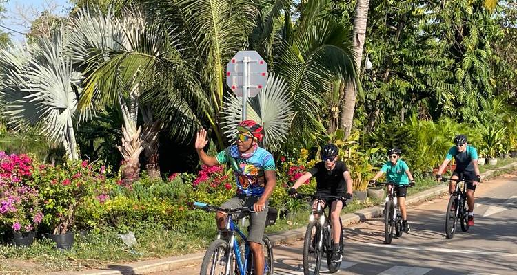 Les cyclistes roulent le long d'une route tropicale bordée de palmiers et de fleurs colorées, un coureur saluant de la main.