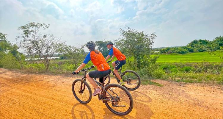 Les cyclistes circulent à vélo tout terrain sur une route en terre rouge à côté de rizières verdoyantes sous un ciel brumeux.