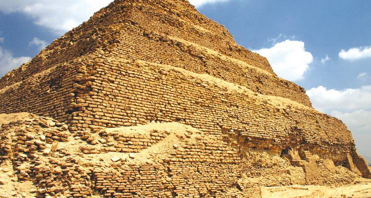 Zijkant van de trapvormige Piramide van Djoser gemaakt van geelachtig steen onder een gedeeltelijk bewolkte hemel.