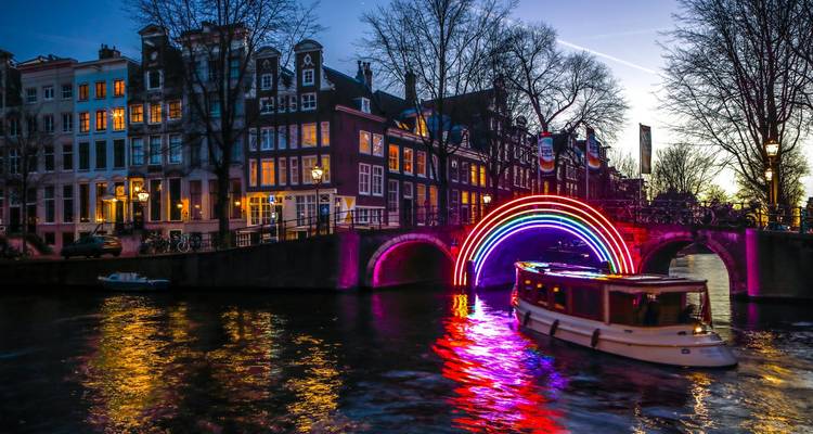 Arco iris colorido de arte lumínico sobre un canal de Ámsterdam con un barco pasando al anochecer.