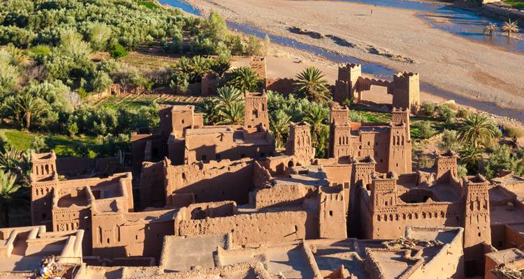 Structures anciennes en terre d'une kasbah regroupées le long d'un lit de rivière asséché avec des palmiers et un paysage désertique en arrière-plan