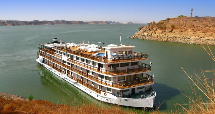 Un navire de croisière blanc à plusieurs ponts sur le Nil glisse sur une eau verte et calme, encadré par des collines arides et un ciel bleu éclatant.