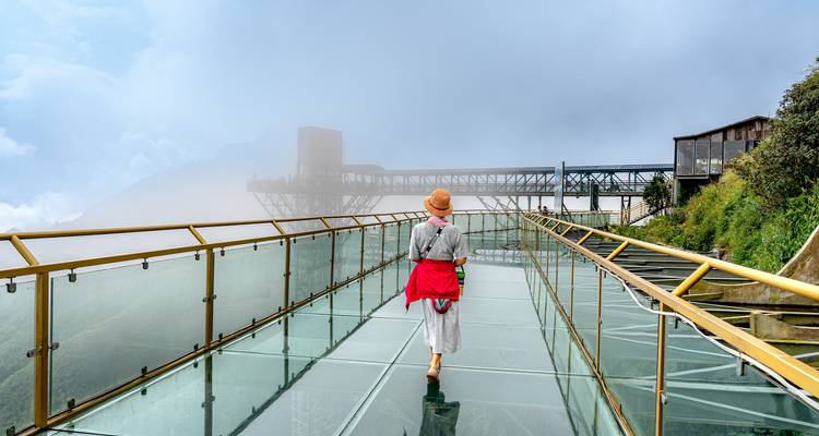 Personne marchant sur une passerelle au sol de verre menant à une structure enveloppée de brouillard.