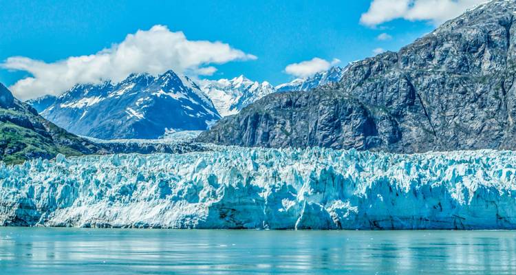 Picos nevados y un enorme glaciar de agua de marea encontrándose con aguas tranquilas del fiordo bajo un cielo azul