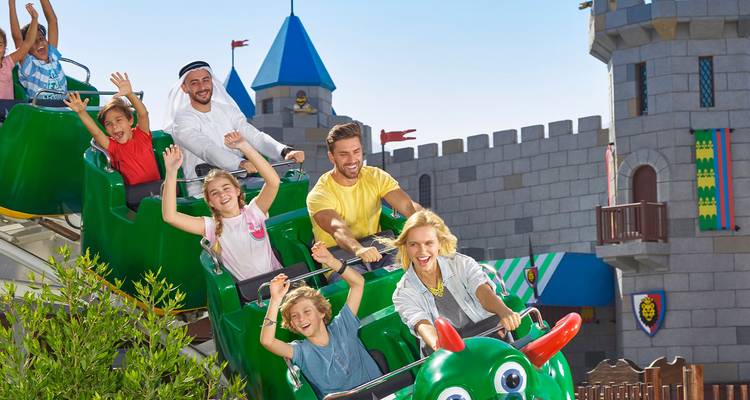 Des familles et des enfants lèvent les bras de joie en faisant un tour sur de petites montagnes russes dans un parc d'attractions à thème.