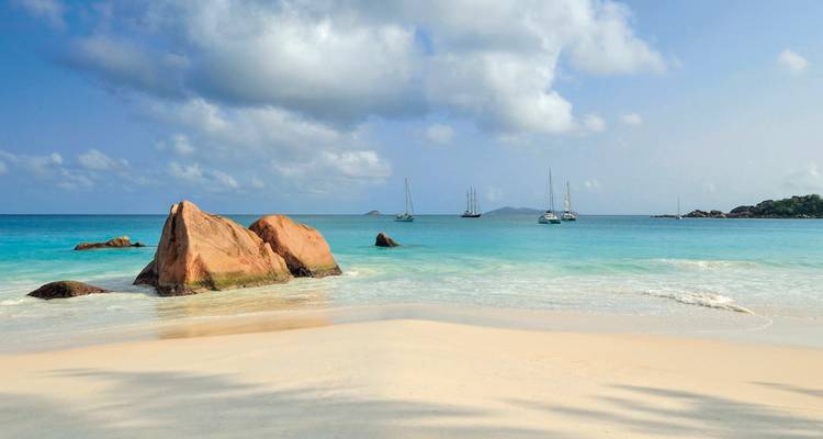 Unberührter weißer Sandstrand mit rosa Granitfelsen und türkisfarbenem Wasser, das mit vor Anker liegenden Segelbooten gesprenkelt ist.