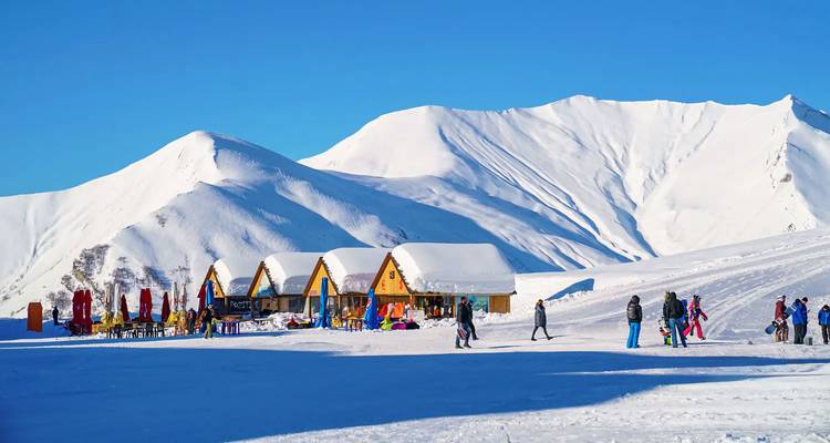 Estación de esquí alpina cubierta de nieve con pequeños chalets de madera y esquiadores moviéndose por una pendiente blanca brillante bajo un cielo azul