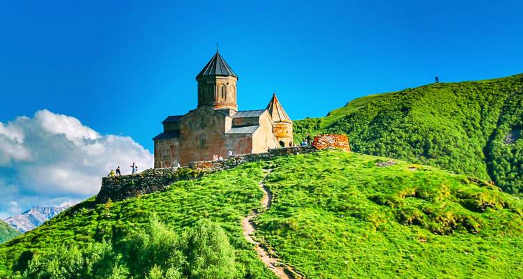 Iglesia de la Trinidad de Gergeti encaramada en una colina verde vívida con un sendero serpenteante que asciende