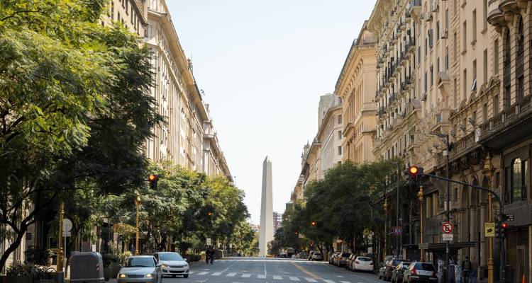 A city street with a view of a distant obelisk, flanked by tall buildings and trees.