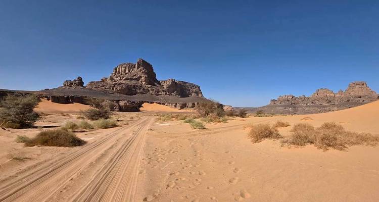 Des traces de véhicule sillonnent un vaste désert encadré par des affleurements rocheux accidentés.