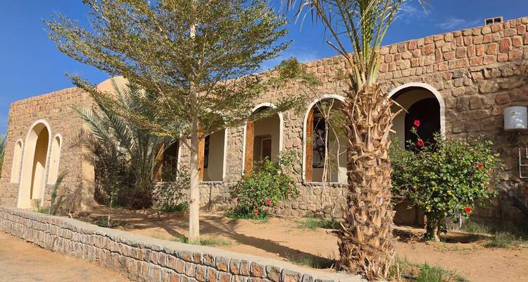 Un pavillon bas en pierre et stuc avec des fenêtres cintrées est ombragé par des palmiers à la lisière du désert.