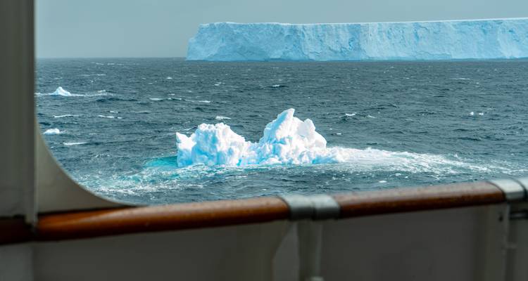 Eisberge treiben auf einem rauen grauen Meer, das von der Schiffsreling aus betrachtet wird, mit einem massiven Tafeleisberg in der Ferne.