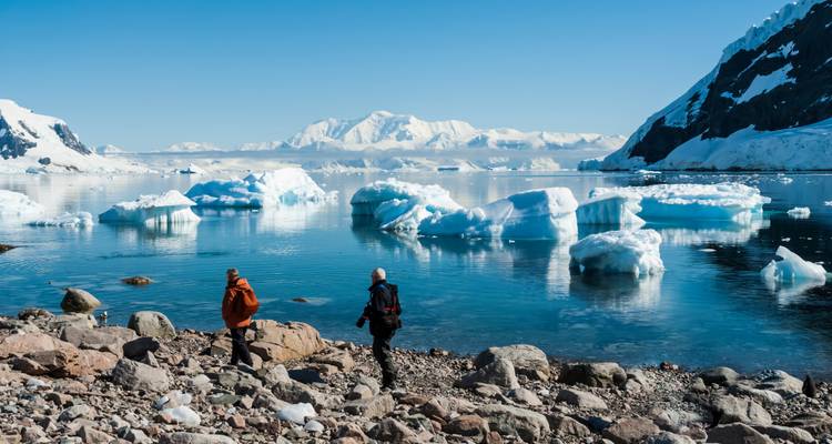 Zwei Expeditionsteilnehmer gehen entlang einer felsigen Antarktis-Küste, wobei Eisberge und verschneite Gipfel sich in ruhigem Wasser spiegeln.