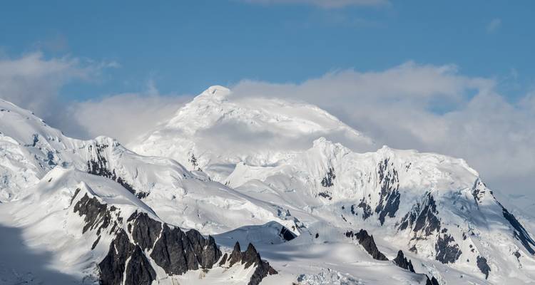 Schneebedeckter Berggipfel erhebt sich über umgebende Gebirgskämme unter blauem Himmel.