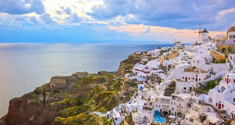 Vista desde el acantilado sobre las casas encaladas de Oia, Santorini, con el Mar Egeo y un cielo dramático al atardecer.