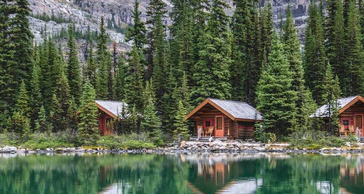 Hölzerne Hütten am Seeufer spiegeln sich in einem türkisfarbenen Alpensee, umrahmt von dichtem Nadelwald