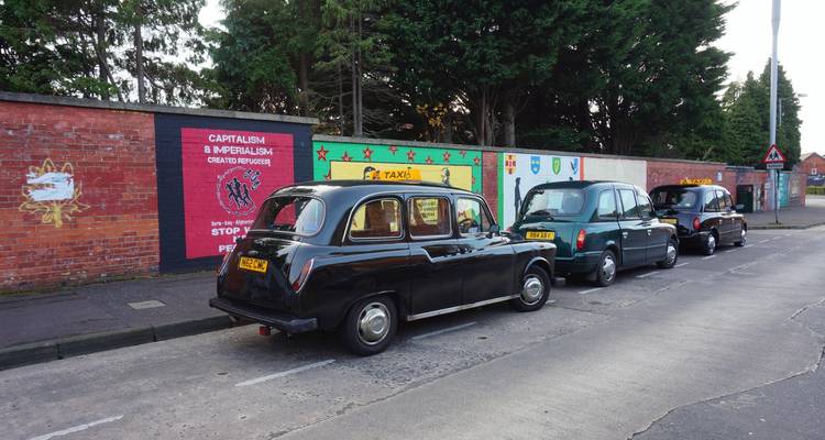 Fila de taxis negros clásicos estacionados junto a murales políticos coloridos en una calle de Belfast.