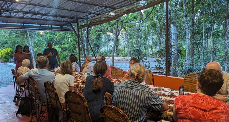 Un gran grupo de viajeros comparte una comida comunitaria en una mesa larga en una terraza de jardín.