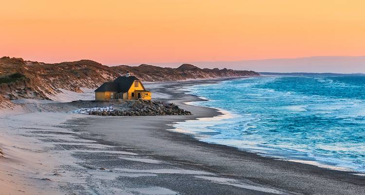 Scène côtière avec une cabane et des dunes au coucher du soleil.