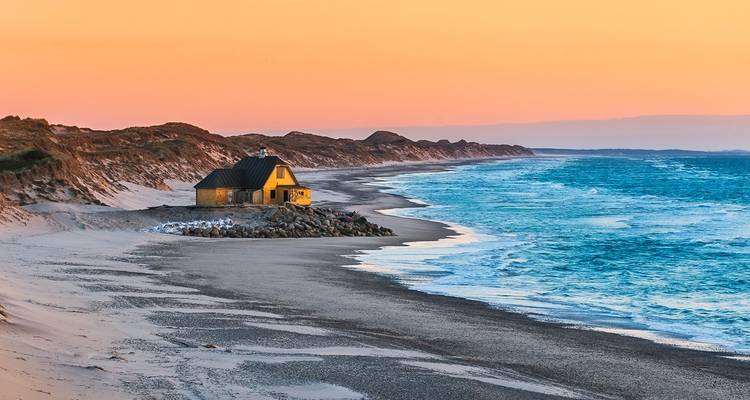 Petite maison jaune isolée située sur une large plage de sable avec des dunes ondulantes et une mer turquoise sous la lumière chaude du coucher de soleil.