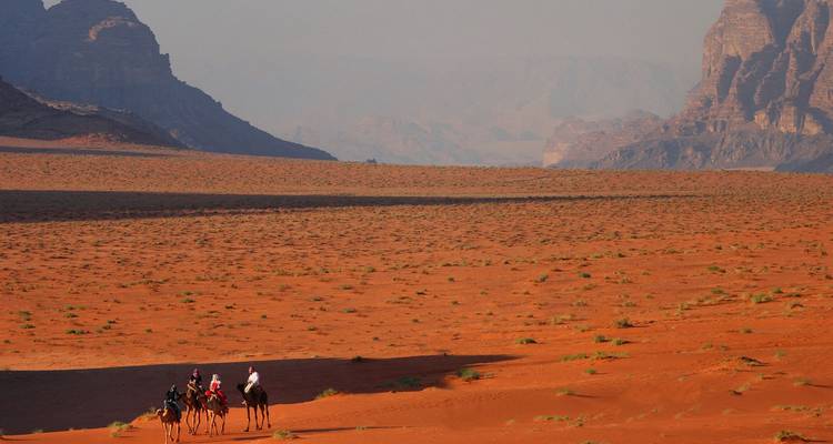 Tres jinetes en camellos cruzan las inmensas arenas rojas y los imponentes acantilados de roca de Wadi Rum.