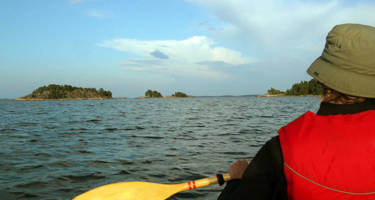 Kajakfahrer mit roter Schwimmweste paddelt über ruhiges Ostseewasser zu kleinen bewaldeten Inseln unter blauem Himmel in Finnland.