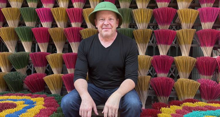 Man in a green hat sits in front of a colorful wall of incense sticks arranged in fan shapes.
