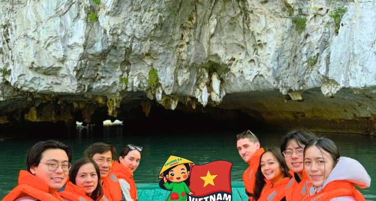 Group in orange life jackets on a boat beneath a limestone cave arch, cartoon Vietnam watermark centered.